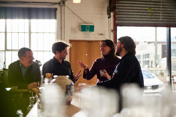 Four people converse around a table in a sunlit room with large windows.