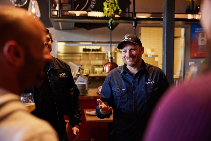 Man in uniform with 'Dave's' logo, smiling indoors with others in casual setting.