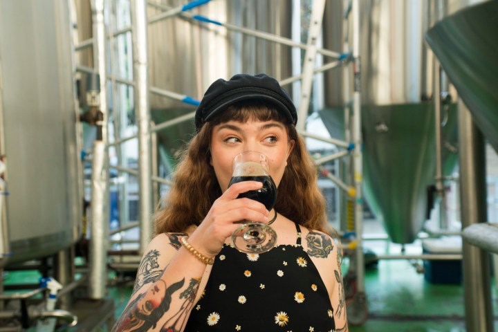 Person drinking dark beverage in brewery with metal tanks.