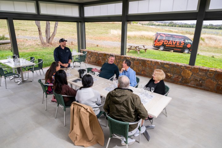 Group of people seated around a table in an outdoor patio with a tour van visible in the background.
