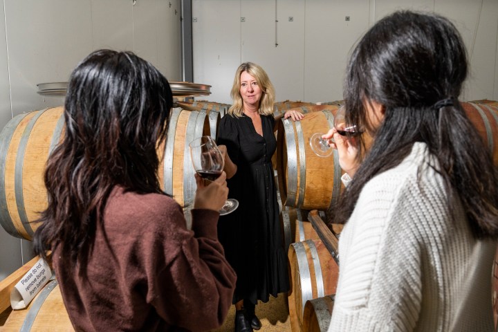 Woman in black dress talks to two people with wine glasses in a barrel room.