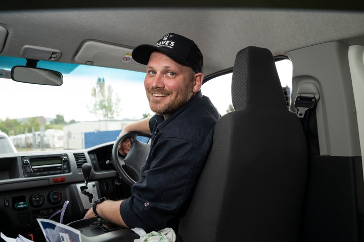 Man in a cap smiling while seated at the driver's seat of a vehicle.