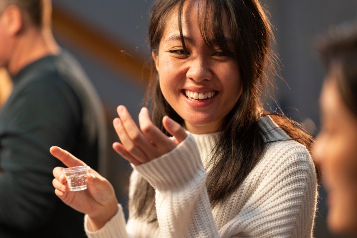 Smiling woman in white sweater holding a small cup and gesturing.
