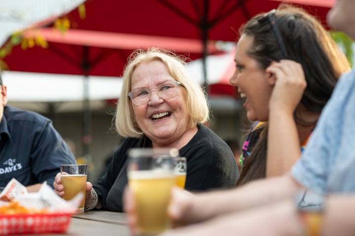 Group of people laughing and drinking beer at an outdoor table with red umbrellas.