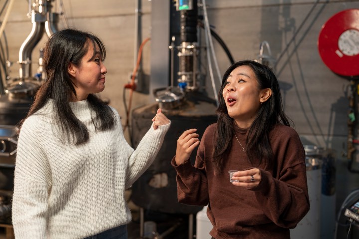 Two women in a distillery talking and smiling, one holding a small cup.