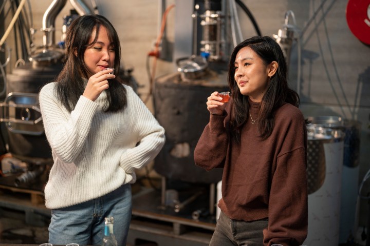 Two women in casual clothing tasting drinks in an industrial setting.